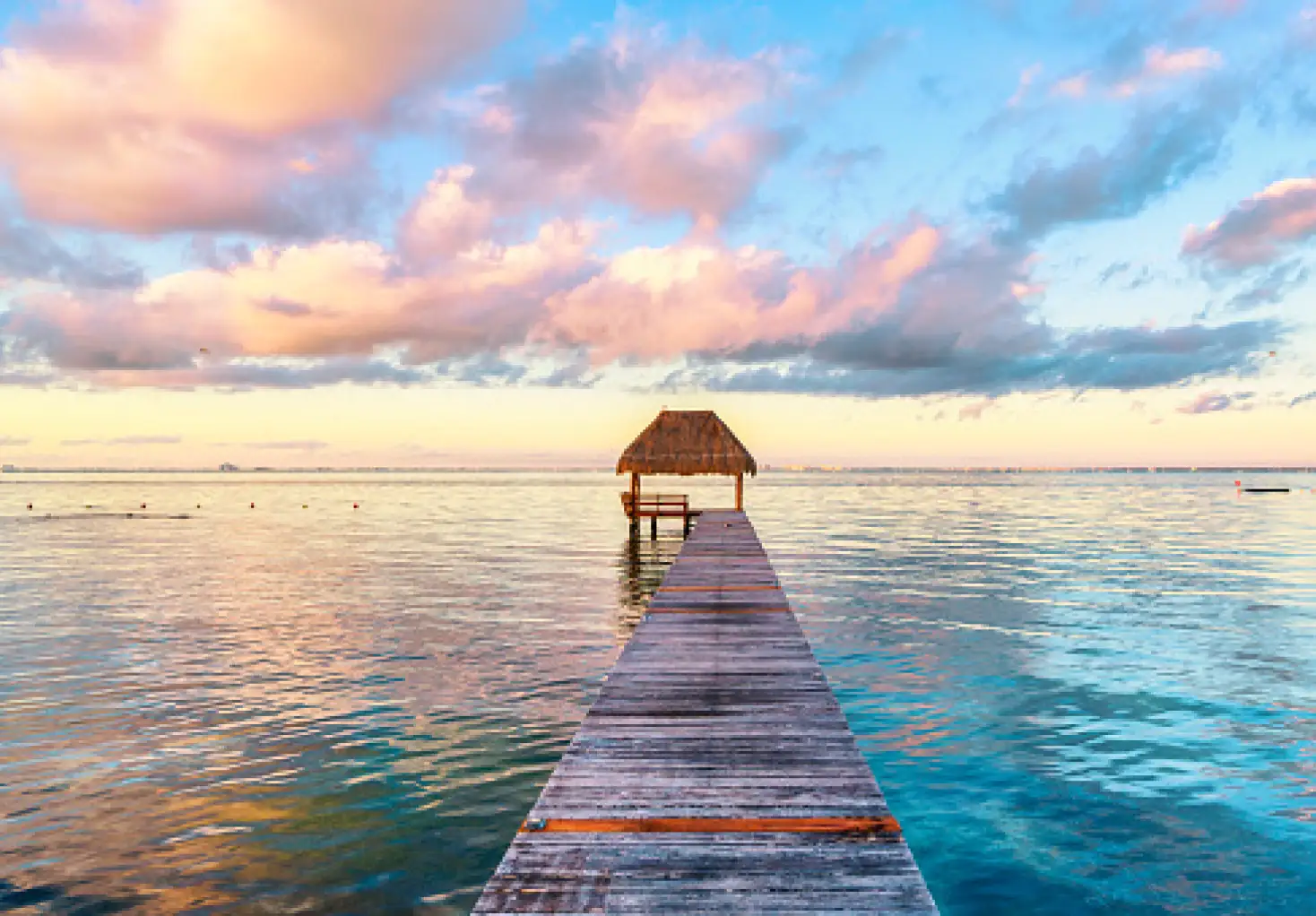 Muelle de madera que se adentra en el mar hacia una palapa sobre el agua, bajo un cielo con nubes en tonos rosados al atardecer.