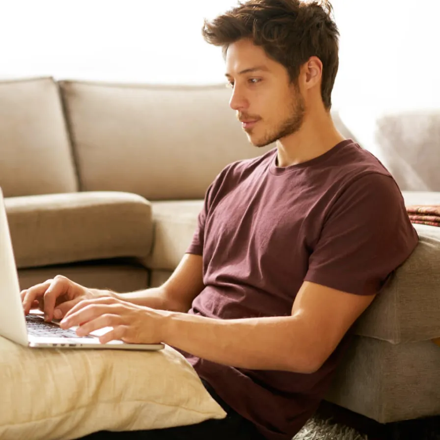 Persona utilizando una computadora portátil desde un sofá en casa, realizando actividades digitales en un entorno residencial cómodo.