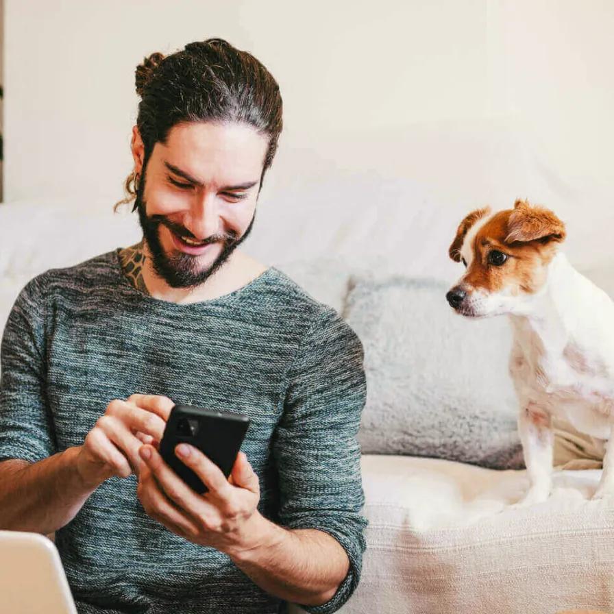 Persona consultando información financiera desde un teléfono móvil en casa, acompañada por su mascota en un entorno residencial.