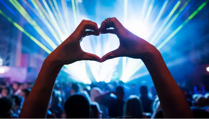 Manos formando un corazón frente a luces de escenario y público en un evento, destacando un ambiente de entretenimiento en vivo.