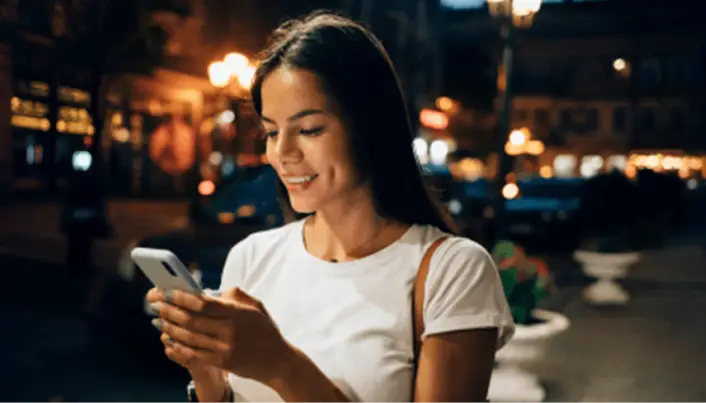Persona utilizando un teléfono móvil en una calle iluminada por la noche, consultando información desde su dispositivo en un entorno urbano.