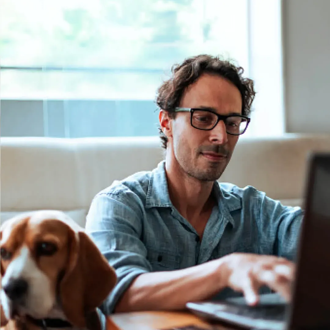 Persona usando una laptop mientras su perro está a su lado en su sala.