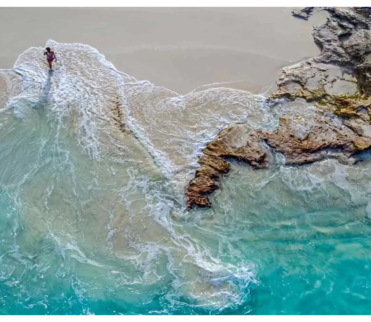 Vista aérea de una persona caminando en la orilla de la playa donde las olas rompen contra rocas.