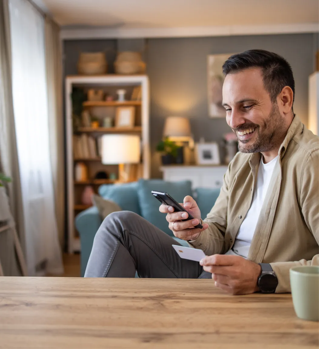 Persona utilizando un teléfono móvil y sosteniendo una tarjeta bancaria desde casa, representando compras en línea, pagos digitales y la gestión segura de finanzas con Banamex.