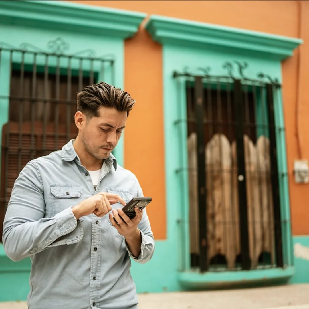 Persona utilizando un teléfono móvil en una calle con fachadas coloridas, representando el acceso a servicios financieros digitales y la gestión de cuentas desde Banamex Móvil.