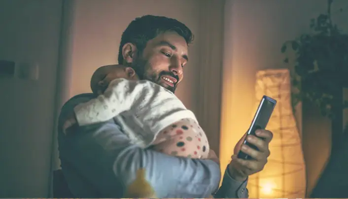 Persona sosteniendo a un menor mientras consulta información desde un teléfono móvil, combinando cuidado familiar y gestión digital.