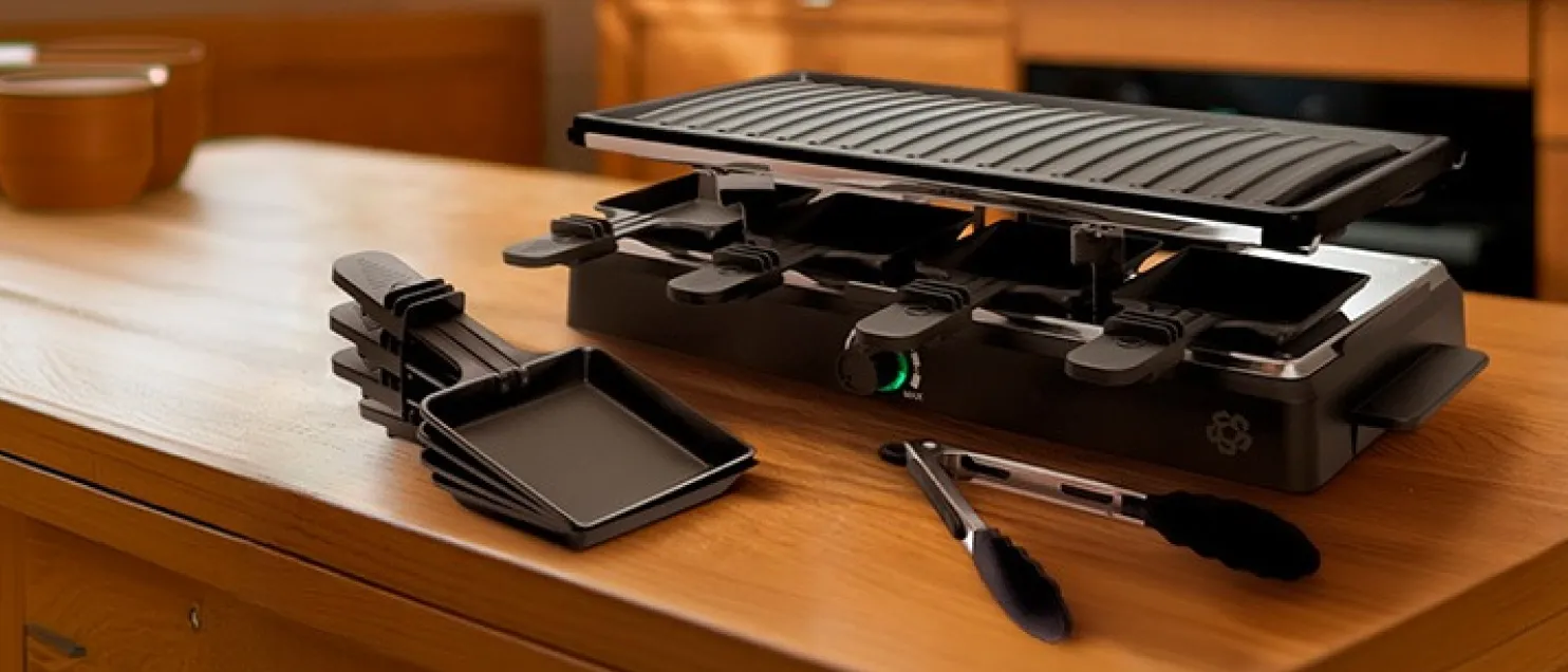 Parrilla eléctrica tipo raclette con bandejas individuales y pinzas, diseñada para preparar alimentos y compartir comidas en reuniones.