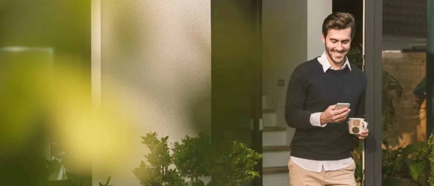Persona consultando un teléfono móvil mientras sostiene una taza, en un entorno residencial con plantas y luz natural.