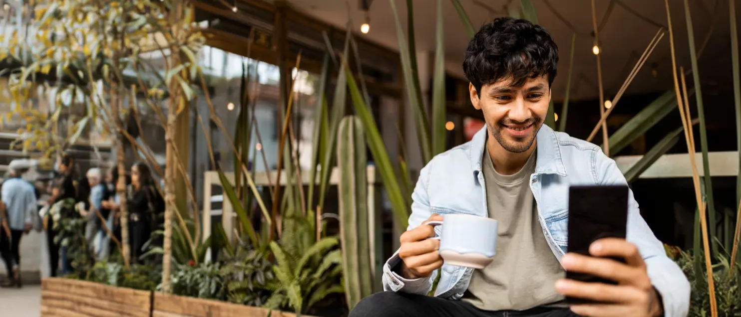 Persona usando un teléfono móvil mientras toma una bebida en una cafetería, rodeada de plantas en un entorno urbano y moderno.