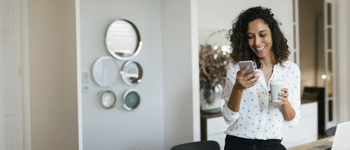 Persona consultando un teléfono móvil mientras sostiene una bebida en un entorno interior moderno, con decoración minimalista al fondo.
