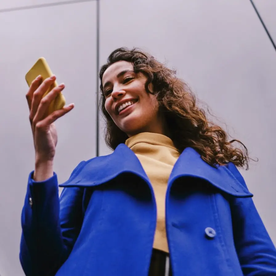 Persona consultando un teléfono móvil al aire libre, con abrigo azul y fondo urbano moderno, mientras revisa información desde su dispositivo.