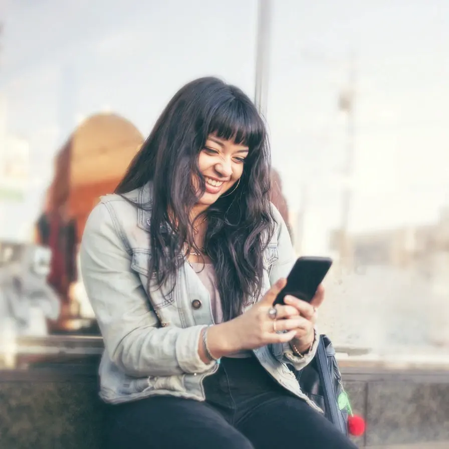 Persona utilizando un teléfono móvil en un entorno urbano, consultando información y gestionando servicios digitales desde cualquier lugar.