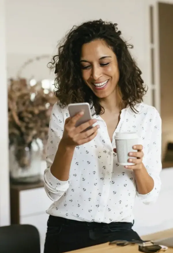 Una mujer sonriendo mientras ve su celular y sostiene un vaso de café.