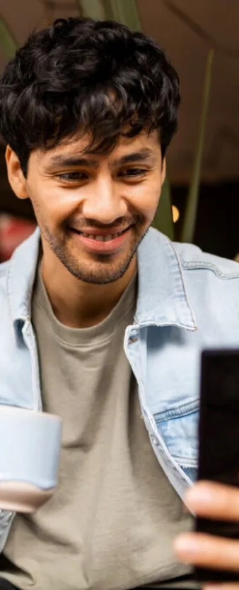 Un hombre sonriendo mientras ve su celular y sostiene una taza de café.