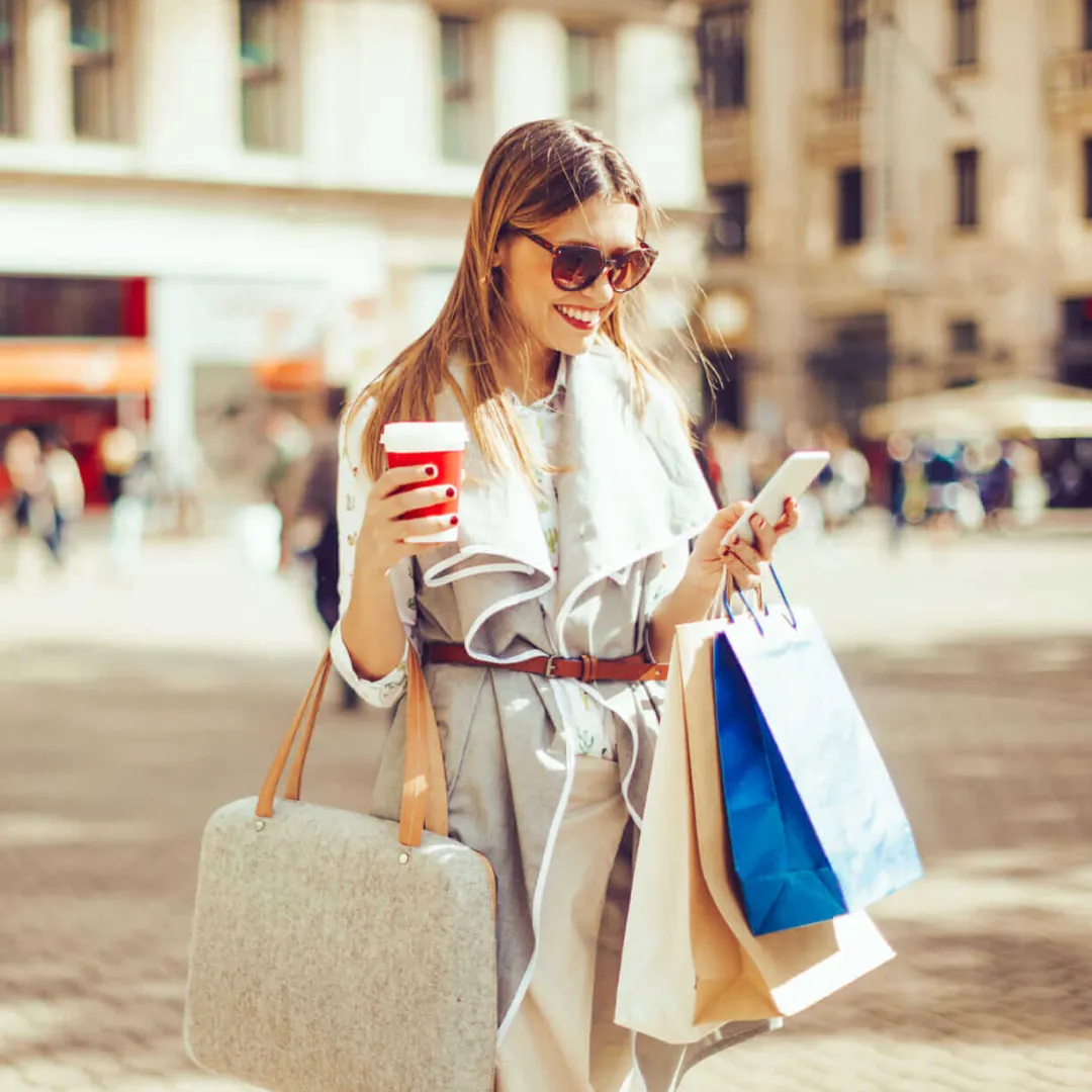 Persona usando un smartphone en un entorno urbano, cargando bolsas de compras y una taza de café.