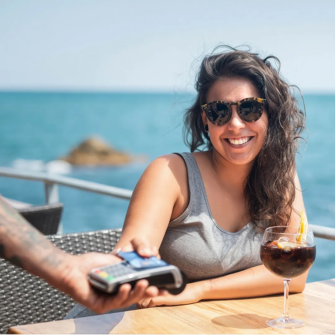 Persona sentada en una terraza junto al mar realizando un pago con tarjeta mediante una terminal bancaria sobre una mesa.