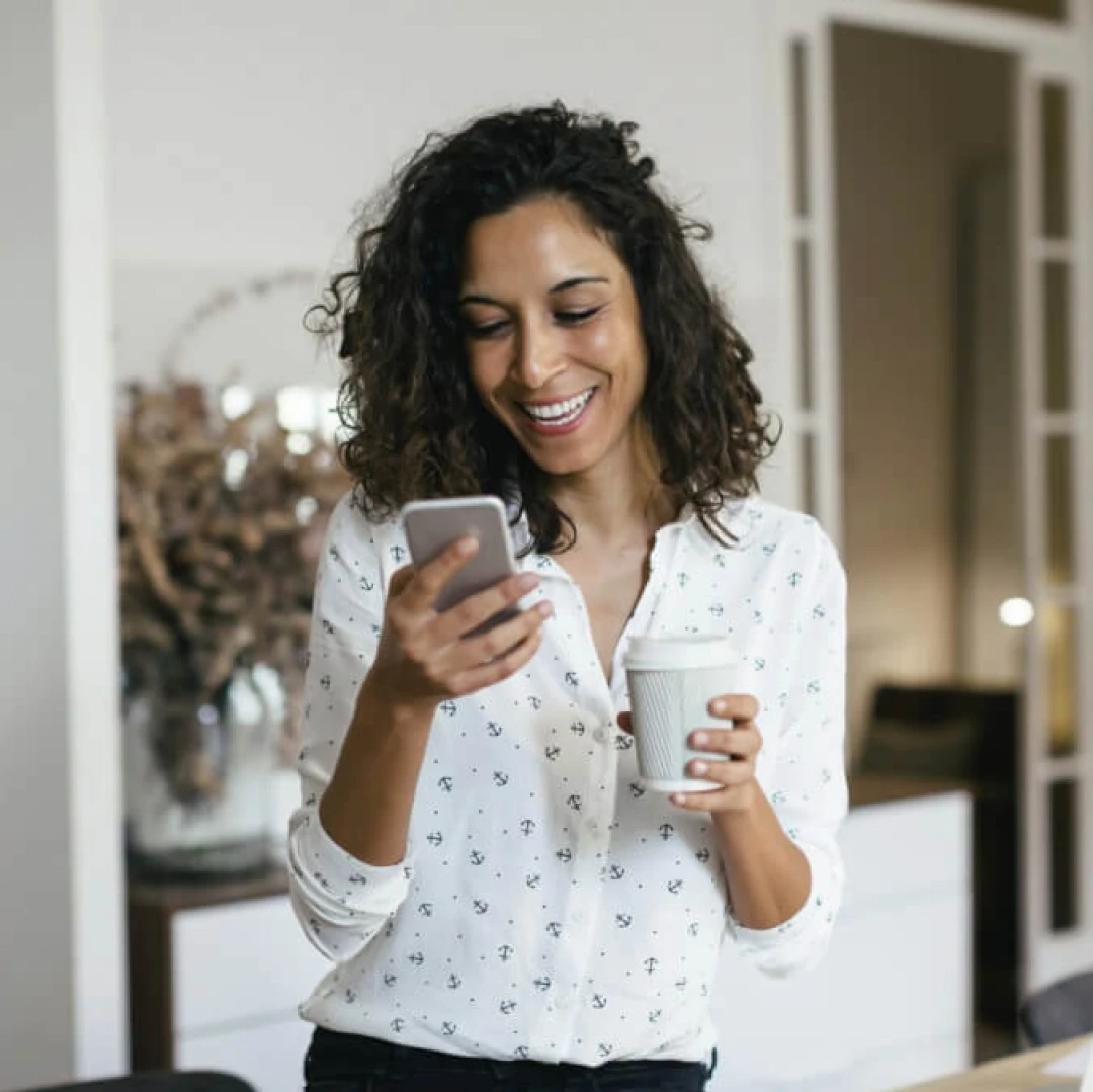 Una mujer sonriendo mientras ve su teléfono y sostiene un vaso de café.