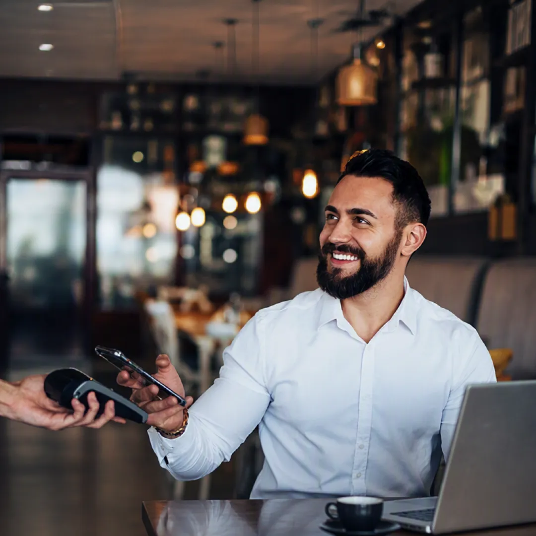 Un hombre sonriendo y pagando la cuenta de un restaurante con su celular.