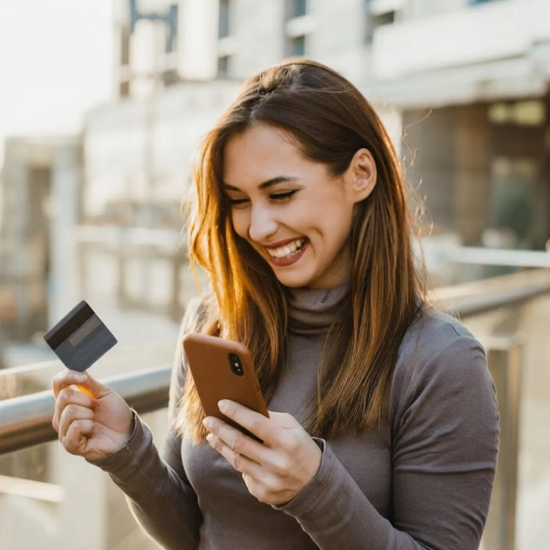 Una mujer sonriendo mientras sostiene su celular y su tarjeta de crédito.