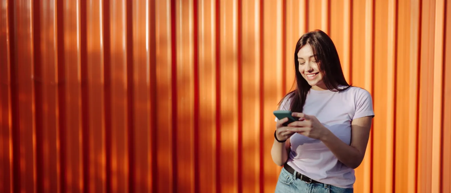 Una mujer revisando su celular frente a un muro naranja.