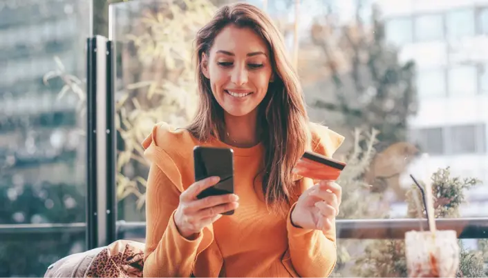Persona realizando una compra en línea desde un teléfono móvil mientras sostiene una tarjeta bancaria para completar el pago digital.