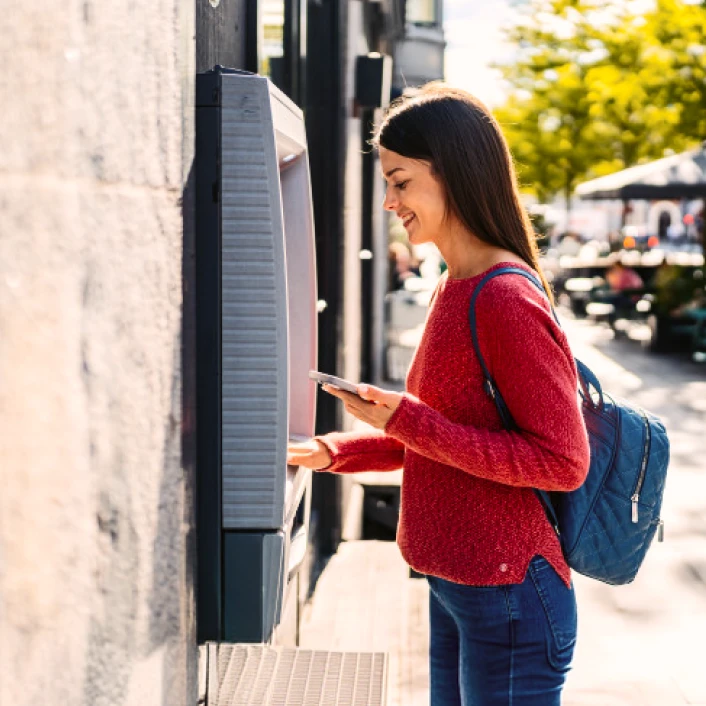 Persona realizando una operación en cajero automático mientras consulta información desde un teléfono móvil en un entorno urbano.