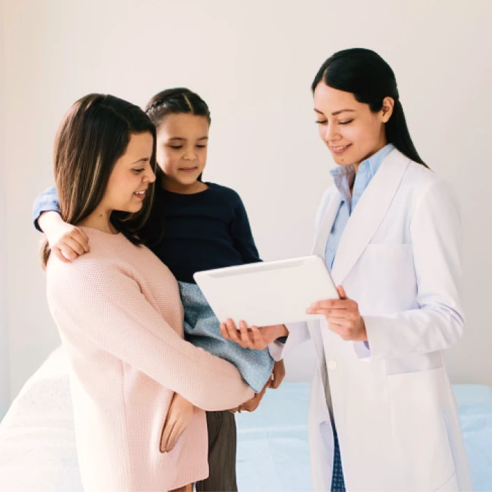 Profesional de la salud explicando información médica a una persona acompañada por un menor durante una consulta de atención preventiva.