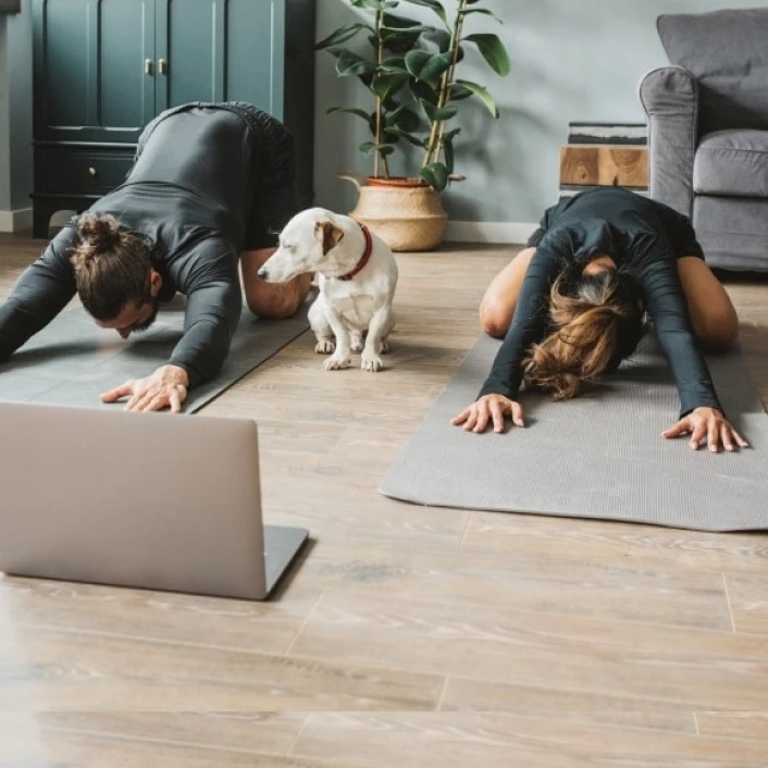 Personas realizando una sesión de ejercicio en casa guiada desde una computadora portátil, acompañadas por su mascota en un ambiente de bienestar.