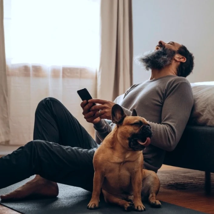 Persona descansando en casa junto a su mascota mientras utiliza un teléfono móvil, reflejando bienestar, tranquilidad y conexión digital.