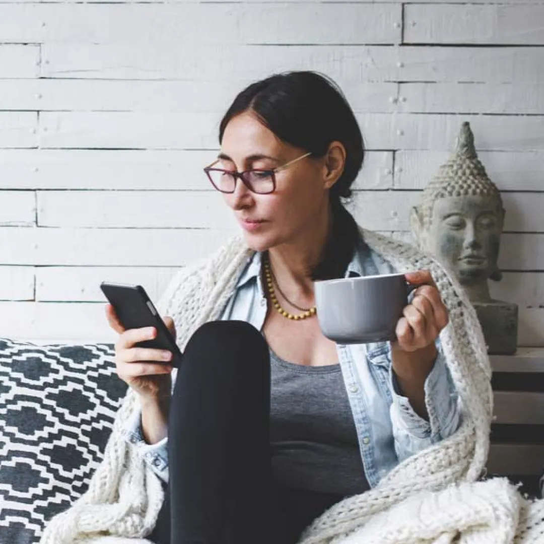 Mujer sentada en un sofá usando su teléfono móvil mientras sostiene una taza de café en casa.