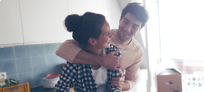 Un hombre abraza a su pareja, una mujer, dentro de una cocina y al fondo se ve una caja de mudanza.