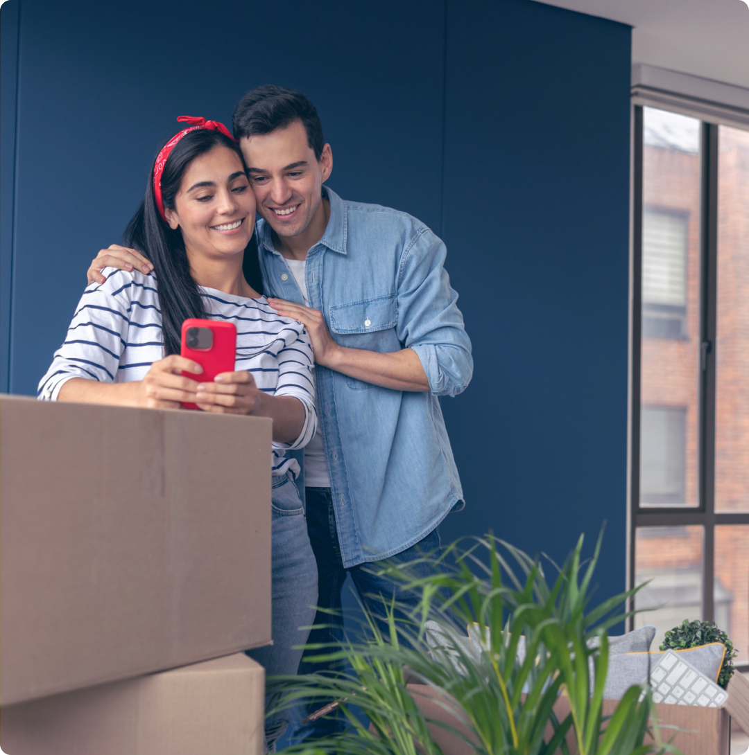 Pareja sonriendo mientras miran el celular de ella, quien está recargada en unas cajas de mudanza.