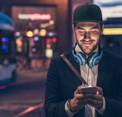 Joven parado en la calle de noche con audífonos colgados en su cuello, sosteniendo y viendo su celular.