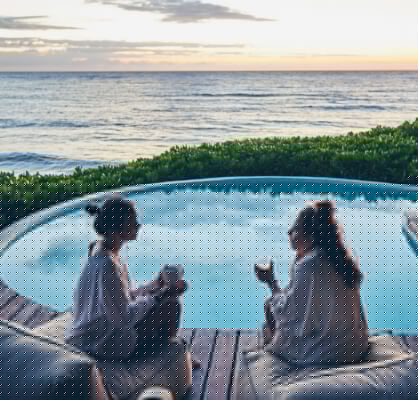 Dos mujeres sentadas frente a una alberca al amanecer, platicando y sosteniendo su taza de café.