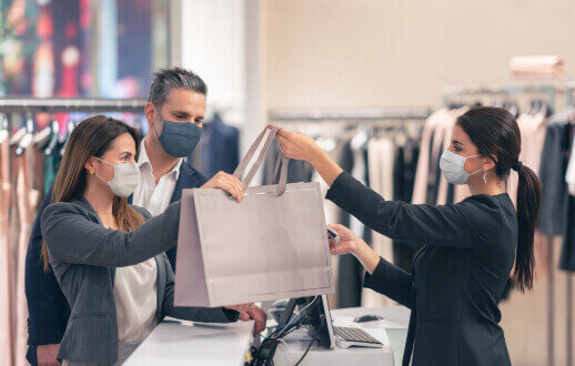 Vendedora entregando una bolsa de compras a una mujer y su esposo, todos usando cubrebocas.