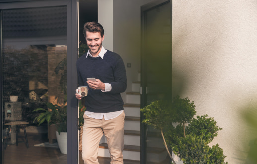 Hombre sosteniendo una taza de café y su celular, haciendo su portabilidad móvil mientras está recargado en una puerta.