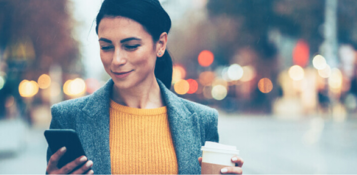 Una mujer sosteniendo su celular y un vaso de café para llevar mientras camina en la calle.