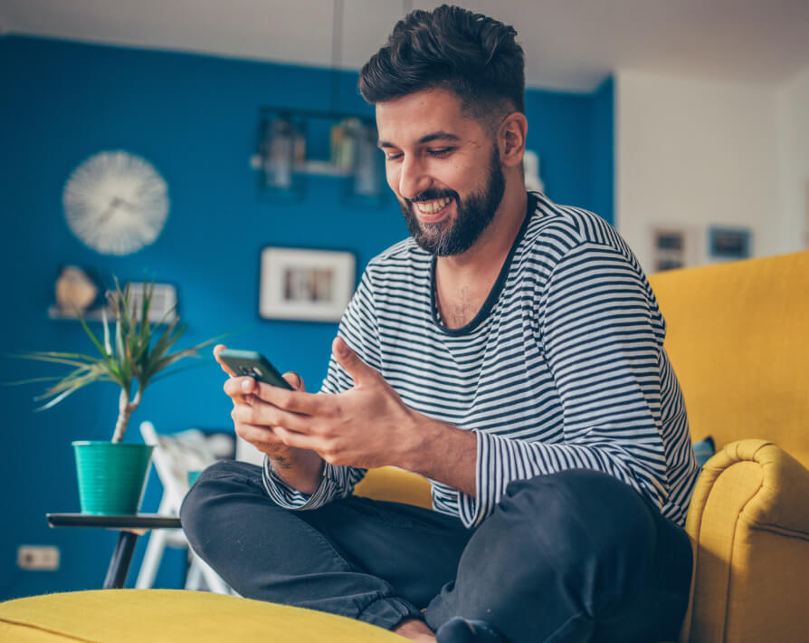 Un hombre sonriendo al mirar su celular en la sala de su casa. 