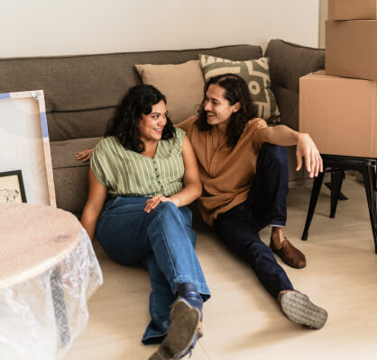 Pareja sentada en el suelo frente a un sillón, con cajas de mudanza a su alrededor.