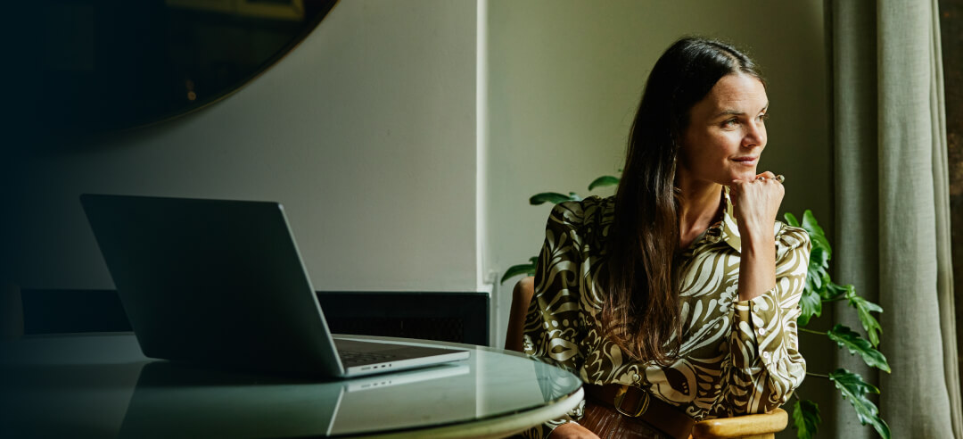Mujer sentada en su escritorio, mirando hacia la ventana mientras reflexional.