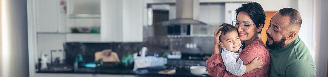 Mujer sosteniendo a su bebé, mientras el papá lo hace reír detrás de ella en la cocina.