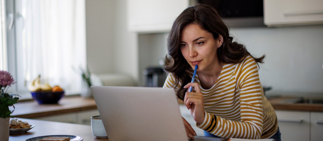 Mujer joven con playera a rayas frente a una computadora con libreta a lado, sostiene una pluma en la mano derecha y la apoya en su barbilla.