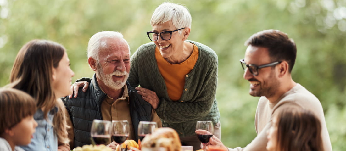 Pareja de adultos mayores sentados en el exterior, rodeados de dos jóvenes y dos niños. Frente a ellos una mesa con frutas, comida y vino.