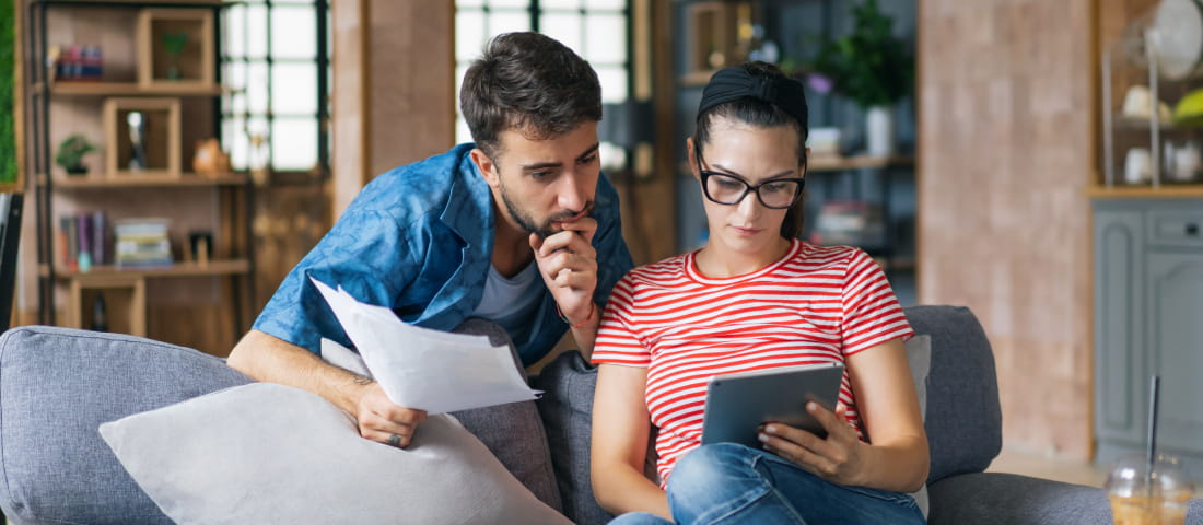 Una pareja, hombre y mujer en su sala revisando papeles e información de créditos en una tablet.
