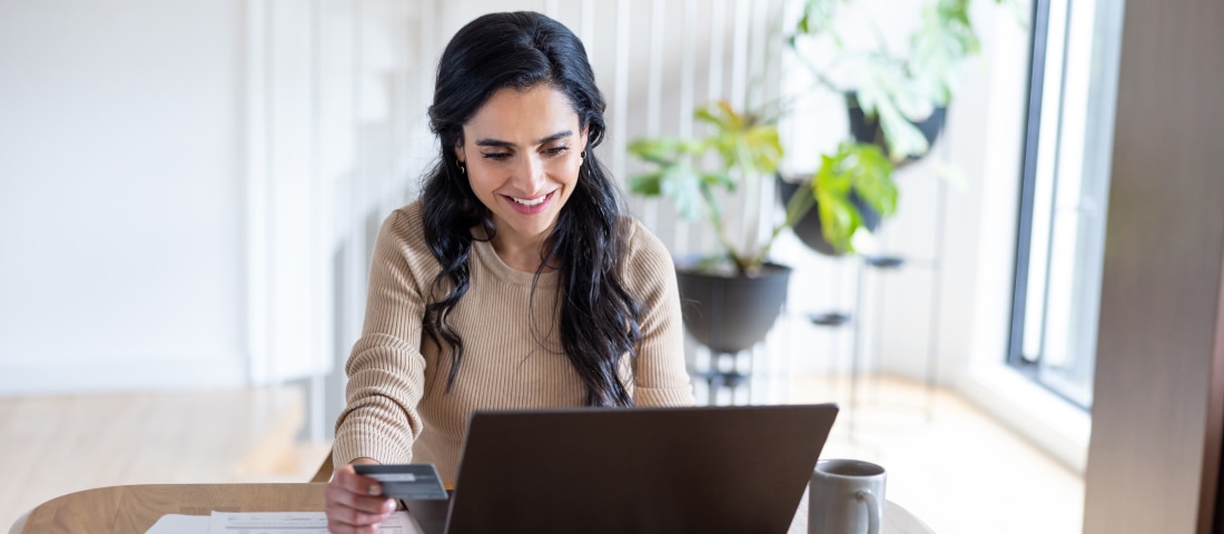 Una mujer sentada frente a su computadora en su sala, con una tarjeta de débito en la mano. 