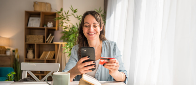 Mujer jóven sentada a la mesa con una taza y un libro frente a ella, sostiene su celular y una tarjeta mientras observa la pantalla de su celular.