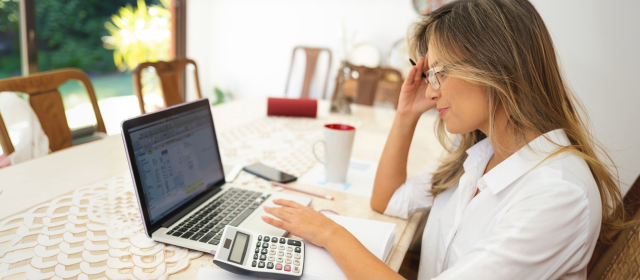 Mujer joven sentada en a sala de su casa revisando sus finanzas en su laptop junto a una taza de café y una calculadora.