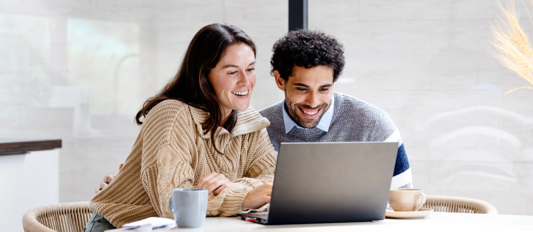 Pareja joven y sonriente, sentados frente a una mesa, señalando la pantalla de la laptop y con una taza de café a un lado.