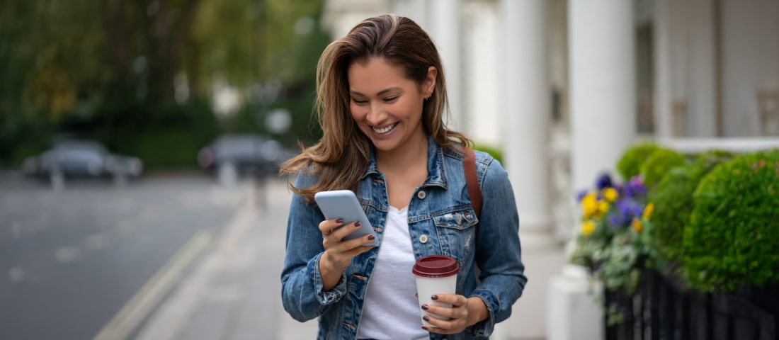 Mujer sonriente revisando su celular con un café en mano, buscando en su celular cómo distribuir su sueldo.