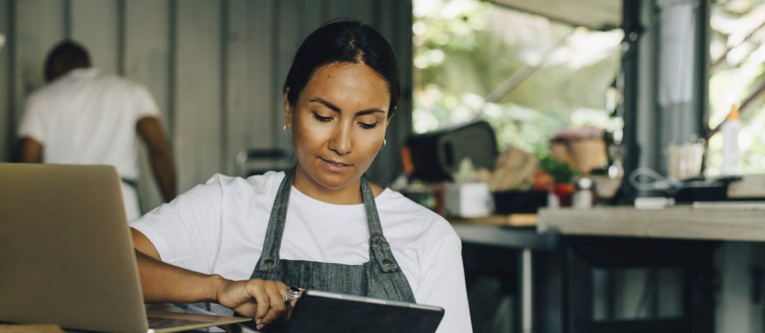 Mujer joven con delantal de pie dentro de un local sosteniendo una tableta en sus manos, frente a ella hay una laptop abierta y otra persona al fondo.